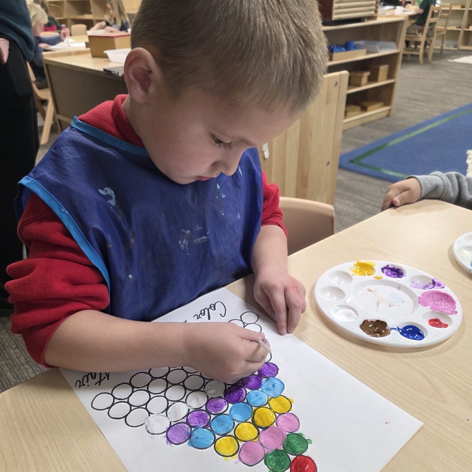 Child painting a colorful design on paper with a paint palette in a classroom setting.